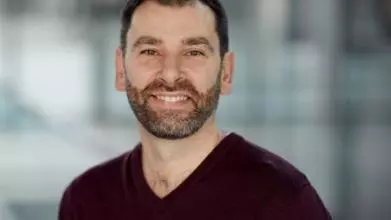 Richard Kemble has short brown hair and a short beard. He is wearing a dark v-neck jumper